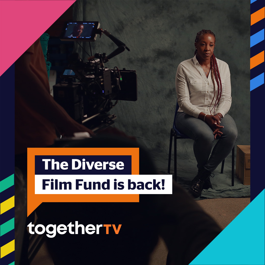 A black woman with long red braid in a white button down, grey pants, and block combat boots, sits on a chair in front of a grey backdrop, looking away from a big film camera that is being operated in the foreground of the photo. Text that appears on the photo reads: The Diverse Film Fund is back! in an orange speech bubble with a logo reading together TV underneath it.