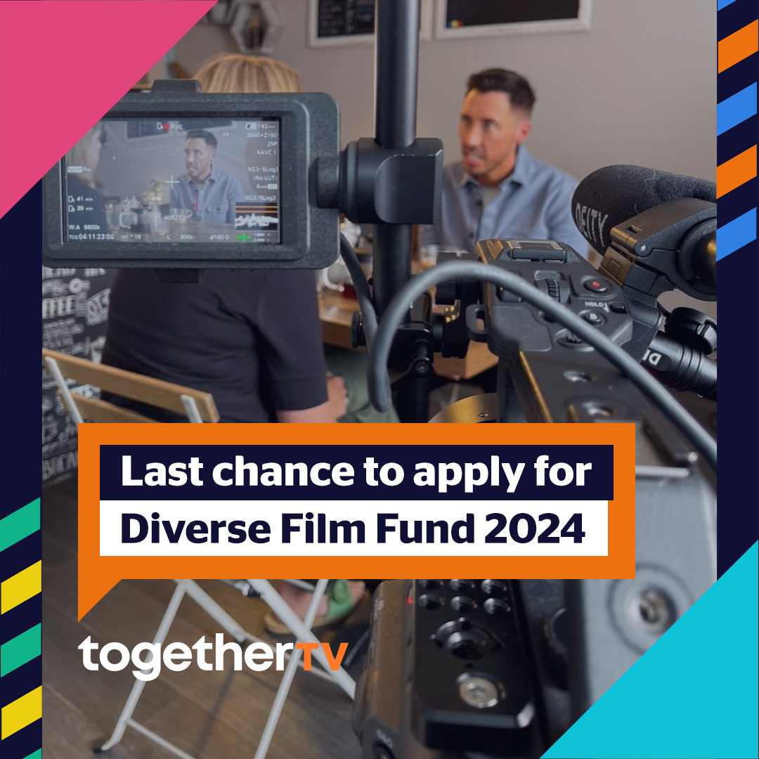 In the foreground there is a big camcorder that is focused on a white man with dark brown hair, in a blue button down shirt who is across the table from a white woman with medium length blonde hair in a black t-shirt, in a café. Text that appears on the photo reads: Last chance to apply for Diverse Film Fund 2024 in an orange speech bubble with a logo reading Together TV underneath it.