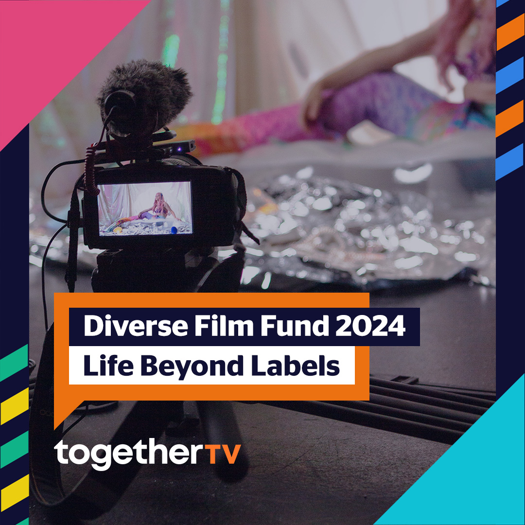 A black camera on a short tripod with a fuzzy microphone on top of it, focused on a woman (mermaid) with pink hair, in a rainbow top and tail. Text that appears on the photo reads: Diverse Film Fund 2024 Life Beyond Labels in an orange speech bubble with a logo reading together TV underneath it.