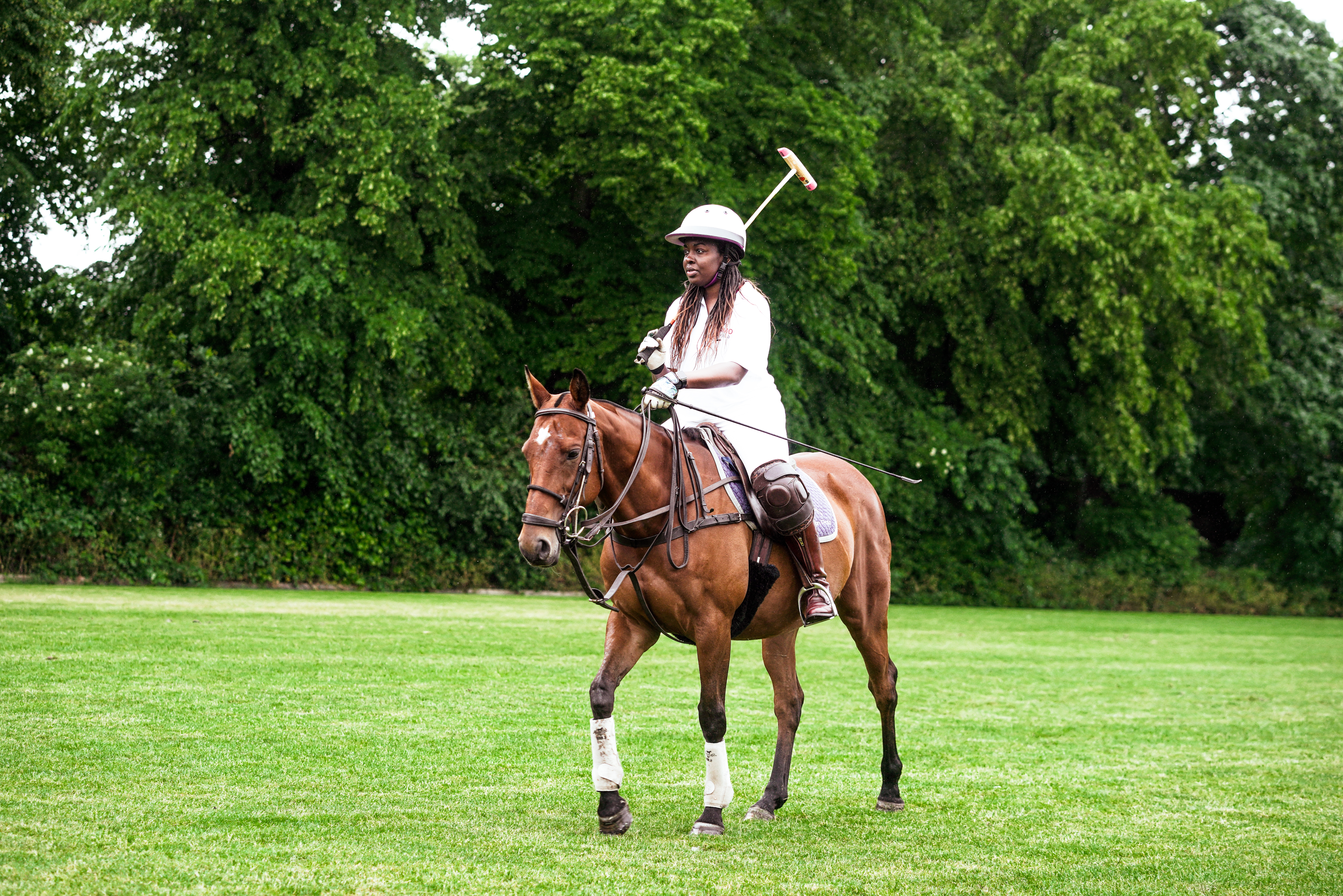 Black female polo equestrian Mame-Yaa Bonsu is sitting on a chestnut coloured horse in polo clothes holding a polo stick trotting on a polo pitch.