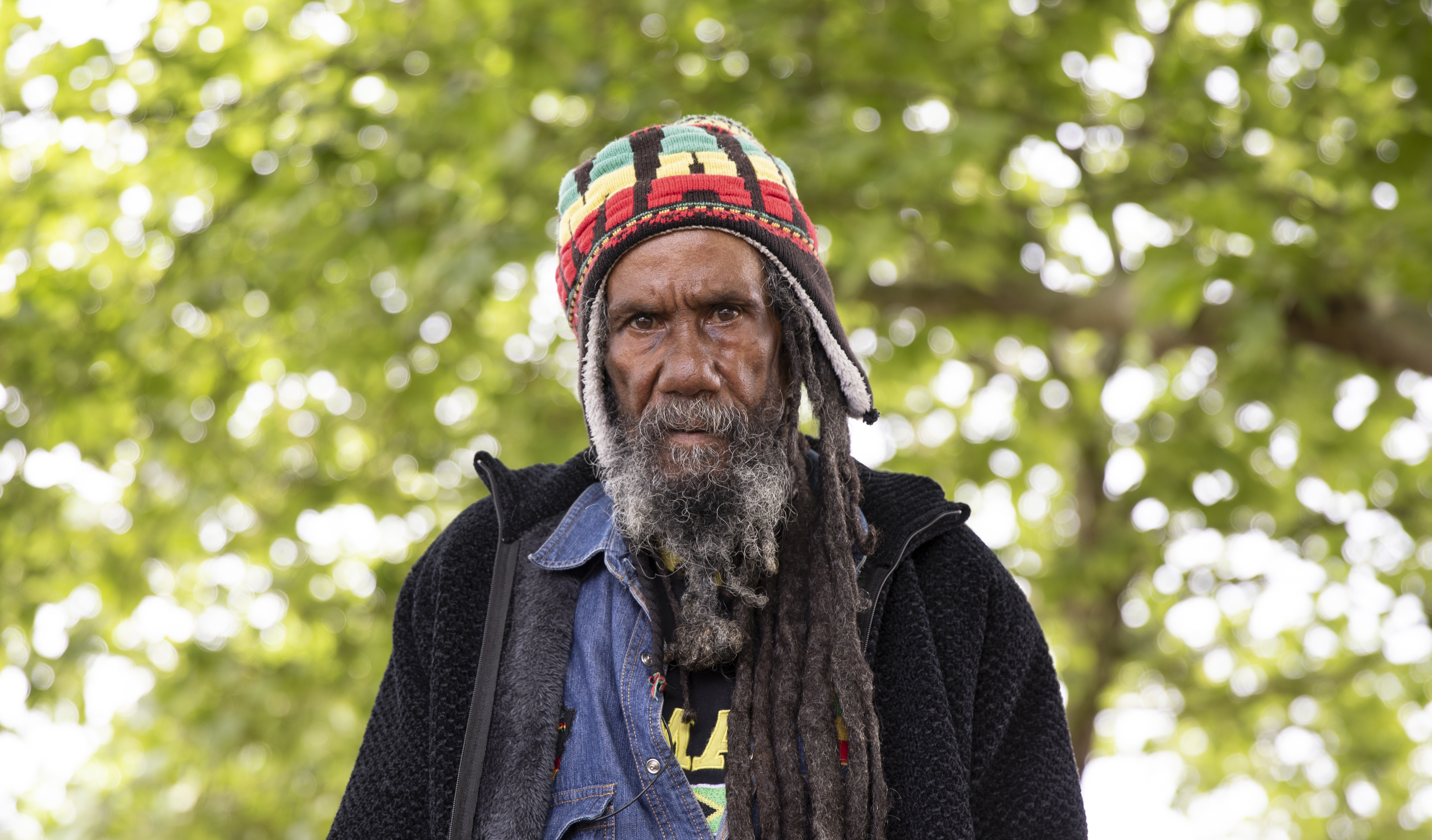 Barry the Beekeeper has long black dreads and a long grey beard wearing a knitted Pan Africa trapper hat and a jean jacket. Behind him are out of focus branches with green leaves.