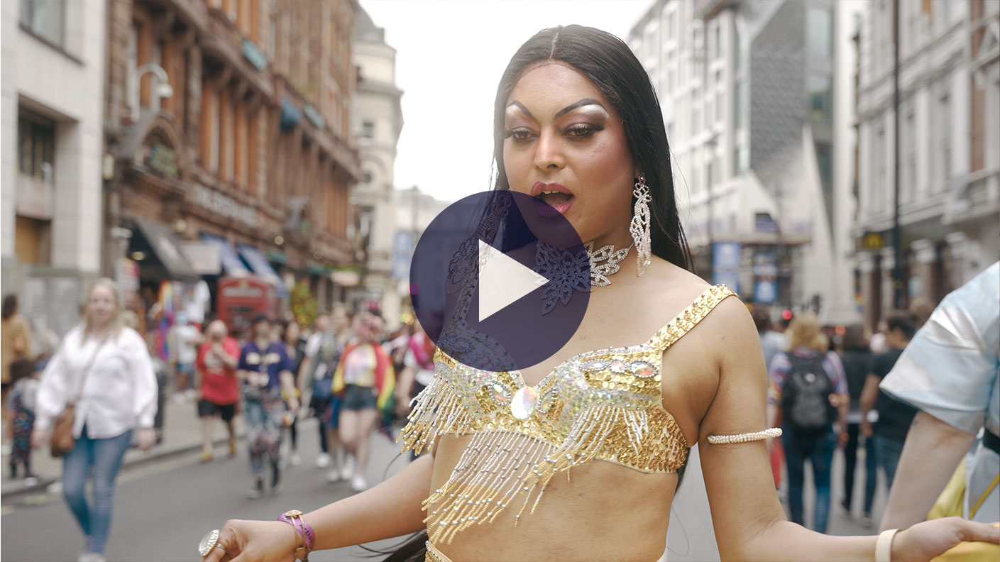 Asifa the muslim, trans drag queen is wearing a sequin and fringe gold bikini top. She has long gold earrings and an elaborate choker necklace on. She has long black hair and dra make up on. She is looking down with her mouth open as she sings and dances. The scene behind her is a street of people walking in both directions lined by London buildings. 