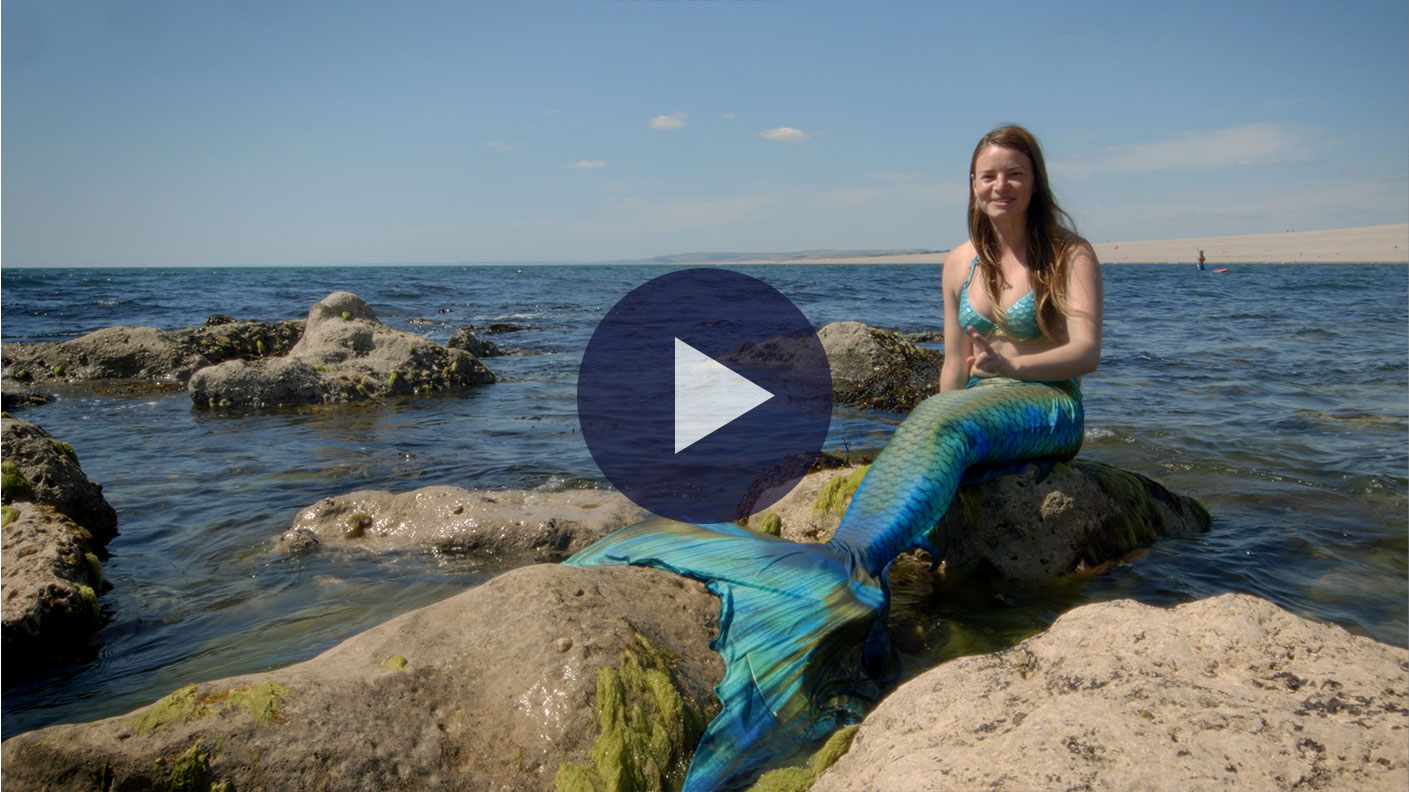 A scene from Mermaids Really Do Exist where a woman dressed as a mermaid is sitting on a rock in the sea wearing a blue and green fin, blue bikini top with long brown hair. Its a sunny day with blue skies.